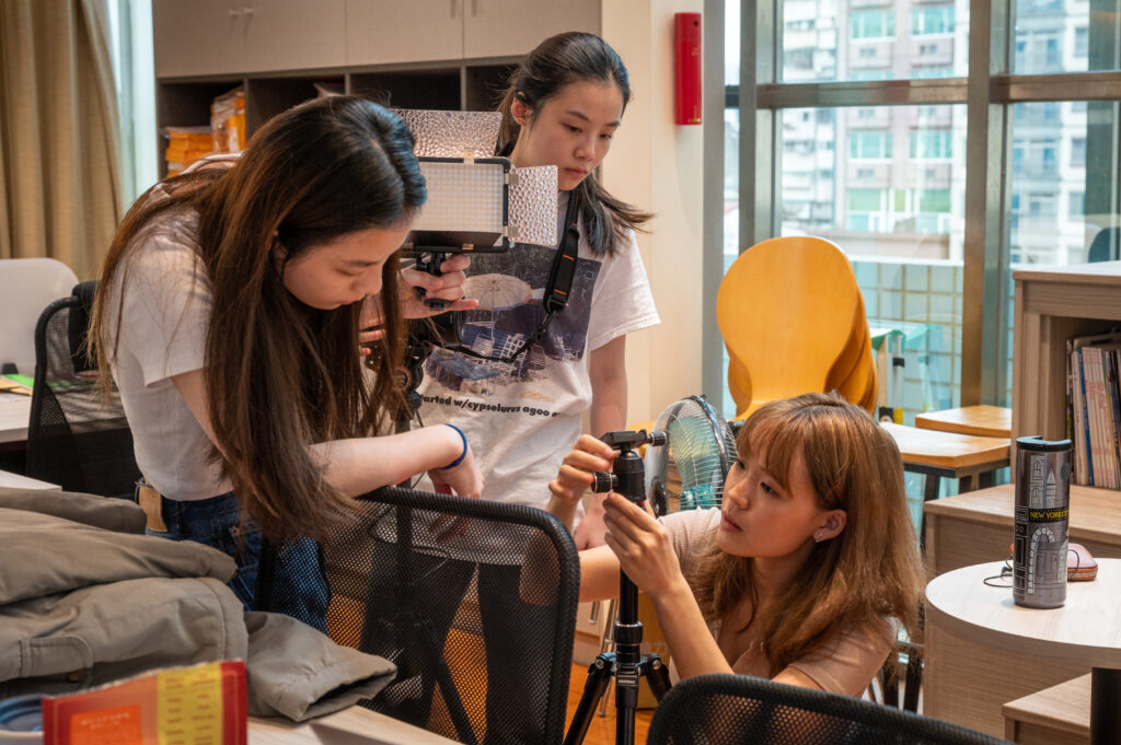 Three crew members one fixing the tripod, one holding a light, and the other person is looking at a tripod.