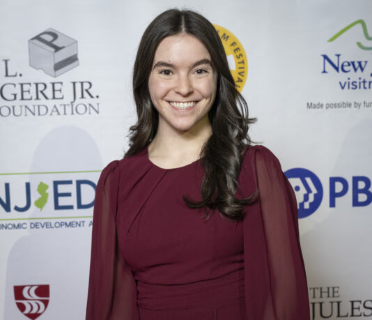 Photograph of Annie Fleisch. She is wearing a long-sleeved maroon dress and smiling. She stands in front of a white wall displaying the logos of various film festival sponsors.