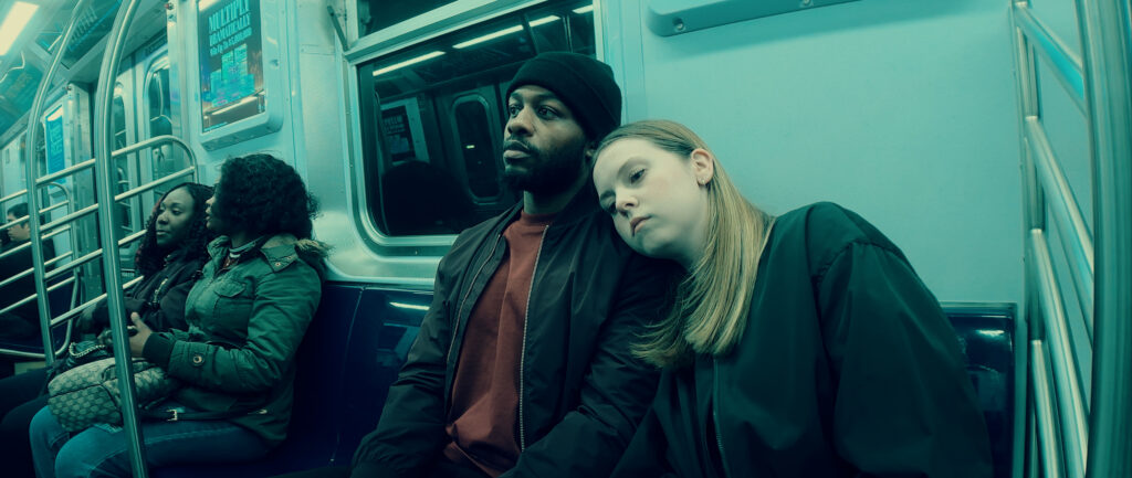 This image depicts a scene inside a subway car. The lighting has a greenish tint, giving the image a moody atmosphere. In the foreground, a person wearing a black hat and jacket is sitting next to a person with blonde hair who appears to be leaning on his shoulder, possibly asleep. Two other passengers are visible in the background.