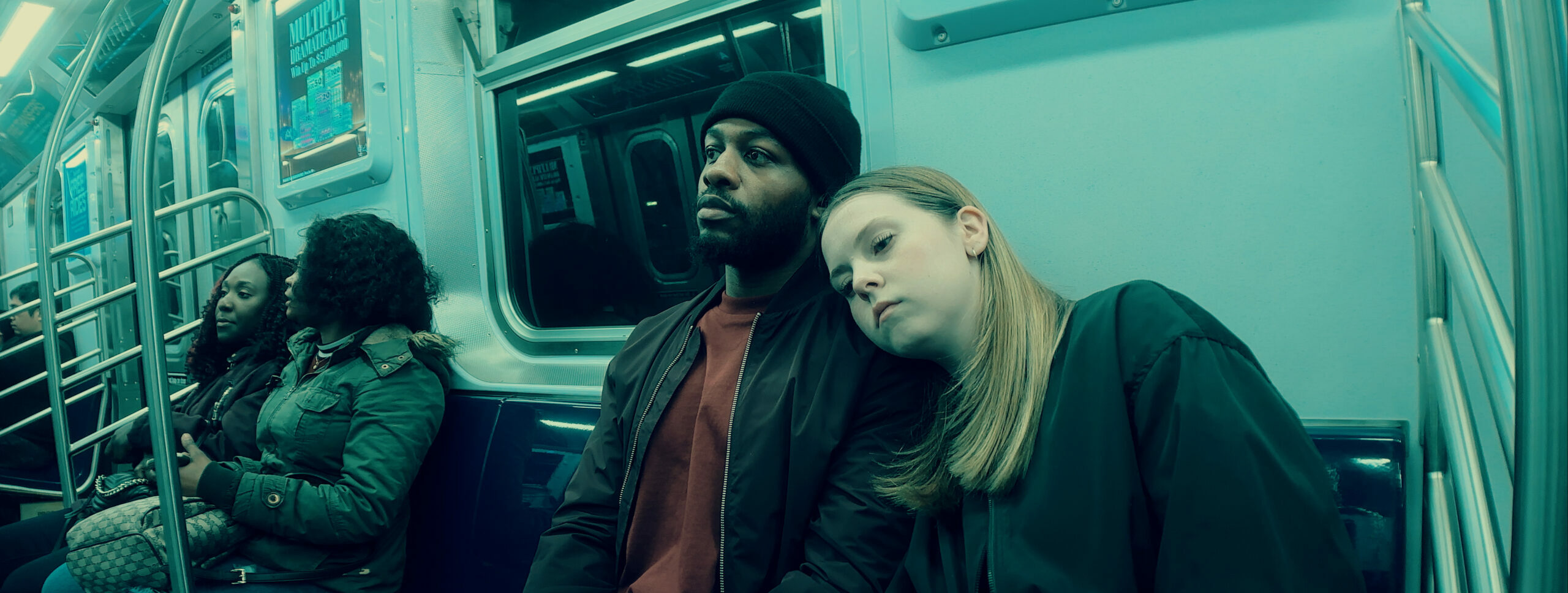 This image depicts a scene inside a subway car. The lighting has a greenish tint, giving the image a moody atmosphere. In the foreground, a person wearing a black hat and jacket is sitting next to a person with blonde hair who appears to be leaning on his shoulder, possibly asleep. Two other passengers are visible in the background.