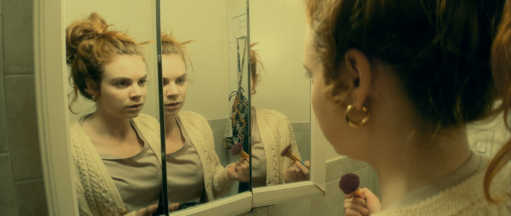 This image shows a woman with blonde hair tied up in a messy bun looking at herself in a bathroom mirror. The mirror is divided into three panels, creating multiple reflections of her face. She's wearing a light-colored cardigan sweater and appears to be applying makeup with a brush. The overall tone of the image has a yellowish tint, giving it a somewhat somber or introspective mood.