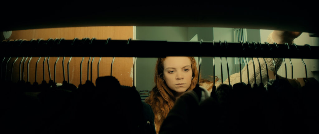 This photo shows a woman with long red hair viewed through a rack of clothes hangers. She appears to be in a closet or clothing store. The woman's face is visible between the hangers, and she has a serious or pensive expression. The image has a cinematic quality, with warm tones and dramatic lighting. 