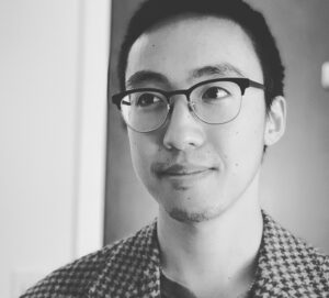 This is a black and white portrait photograph of Directot Justin Ho, with short dark hair. He's wearing glasses with dark frames and is looking slightly to the side with a subtle smile. He's wearing a patterned collared shirt.