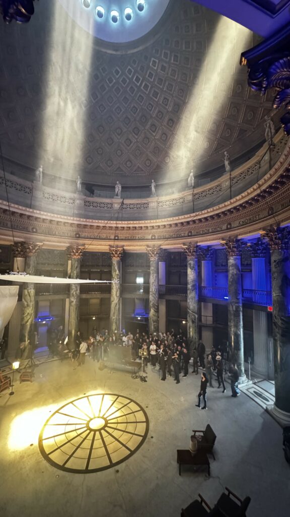 A group of people behind a TV connect to the camera watching as the DP shows them the camera choreography they will do for the scene. There’s a 20X20 frame hung close to the ceiling. Two 10K lights face the floor, creating an angelic look in the space.