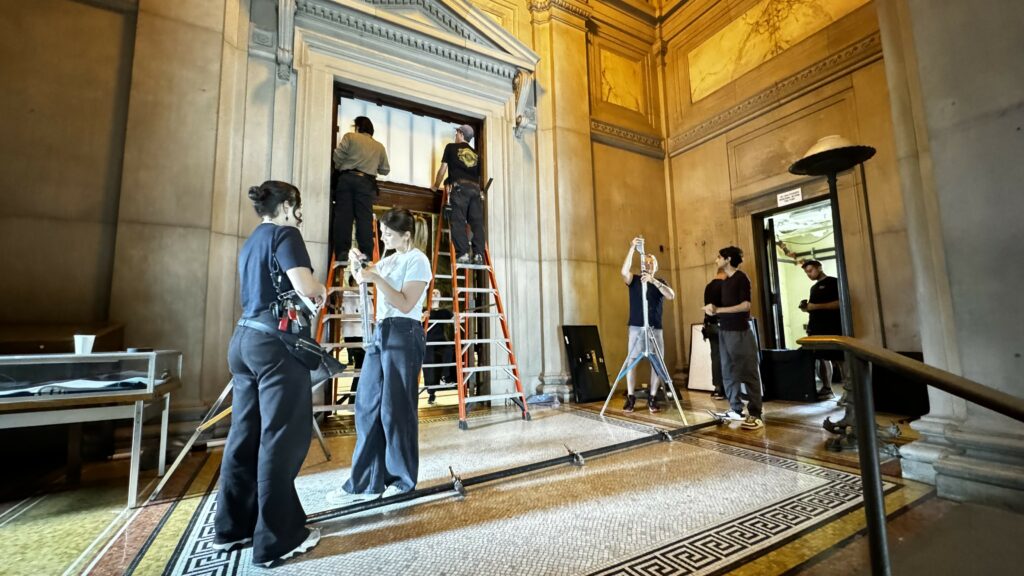 Two girls are standing by a ladder setting up a rig. Behind them, two men stand on top of two ladders, tapping defusion to the window. To this right, one man is setting up a C-Stand while the other is watching.