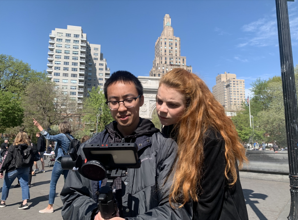 This photo is taken in what appears to be Washington Square Park in New York City. In the foreground, two people are looking at a camera or filming device. One is Director Justin Ho, wearing glasses and a gray jacket, while the other is a woman with long red hair. Behind them, the iconic Washington Square Arch is visible, along with several tall buildings typical of New York City architecture.