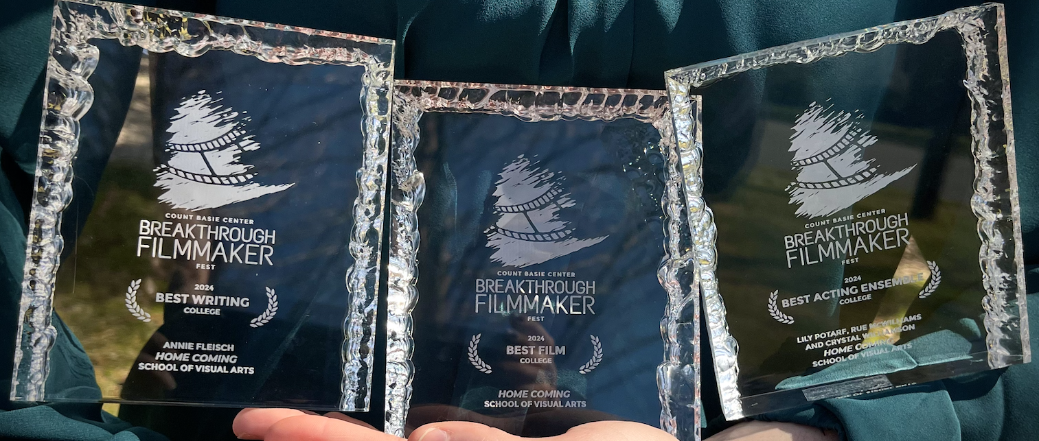 Photograph of a pair of arms holding three square glass plaques. All three plaques feature white engraved text displaying the words “Count Basie Center Breakthrough Fest”; the bottom text of each plaque describes a different award. Left plaque: “2024 Best Writing College; Annie Fleisch; Home Coming; School of Visual Arts.” Center plaque: “2024 Best Film College; Home Coming; School of Visual Arts. Right plaque: “2024 Best Acting Ensemble College; Lily Potarf, Rue McWilliams; Crystal Williamson.”