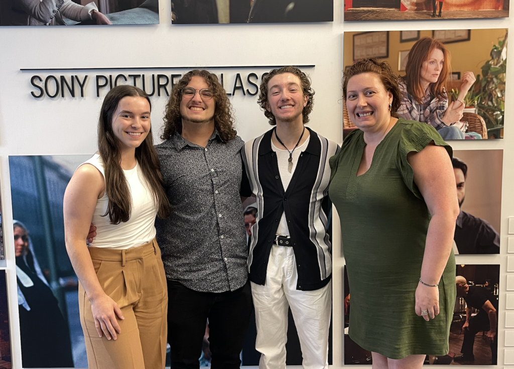 Photograph of four people smiling in front of a white wall. The wall behind them is labeled “Sony Pictures Classics” in big black text and displays printed still images from various movies. 